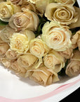 A close-up of a bouquet of pale cream to beige roses wrapped in white paper with a light pink ribbon.