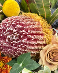 A close-up of a floral arrangement featuring a large pink and white banksia flower, a yellow Billy Button, a beige rose, and orange chrysanthemum, surrounded by green leaves.