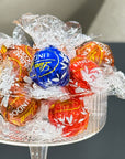 A clear glass bowl filled with assorted wrapped chocolates, including a prominent blue Lindt ball and several red, orange, and gold wrapped candies, photographed close-up against a dark gray background.