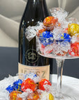 A bottle of Jansz premium cuvée sparkling wine with a clear glass pedestal bowl filled with wrapped chocolates in red, blue, yellow, and orange, Lindt Lindor with more chocolates scattered on the table in front.