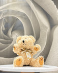 A small beige teddy bear plush toy with a gold ribbon bow sits centered on a round white pedestal against a soft gray rose-shaped backdrop.
