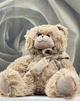A plush teddy bear in a warm beige/tan color with a matching fabric bow, sitting on a white round pedestal against a soft gray rose-shaped backdrop.