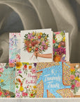A stack of Mini La La Land floral greeting cards displayed against a draped gray fabric background. The cards feature colorful watercolor and illustration designs, including bouquets, birds, butterflies, and celebratory messages such as 'Happy Birthday' and 'A Thousand Thanks'.