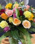 A vibrant rose bouquet featuring a mix of pastel and bright roses, yellow and orange blossoms, pink flowers, and green foliage, arranged in a mason jar container on a wooden surface.