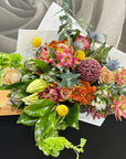 A vibrant floral arrangement featuring a mix of green leaves, gum foliage, banksia, leucodendron and orange flowers, yellow pom-pom billy buttons, and purple thistle-like blooms, styled on a wooden bench with white wrapping against a gray background.