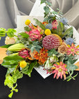 A vibrant wildflower bouquet with banksia, golden Billy Button, and green foliage, wrapped in white paper and resting on a wooden surface.