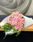 A bouquet of pink roses wrapped in white paper with green foliage, resting on a wooden surface against a dark background.