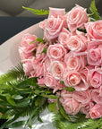 A bouquet of light spray pink roses with green foliage, wrapped in white paper and photographed close up.