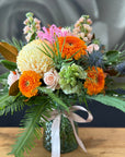 A vibrant flower arrangement in a textured green glass vase, featuring bright orange gerbera daisies, pale yellow chrysanthemums, pink roses, and green foliage, tied with a white ribbon on a wooden surface.