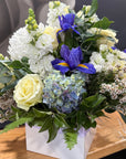 A square floral arrangement in a white box, featuring white and pale yellow roses, blue hydrangeas, purple irises, and green foliage on a wooden surface.