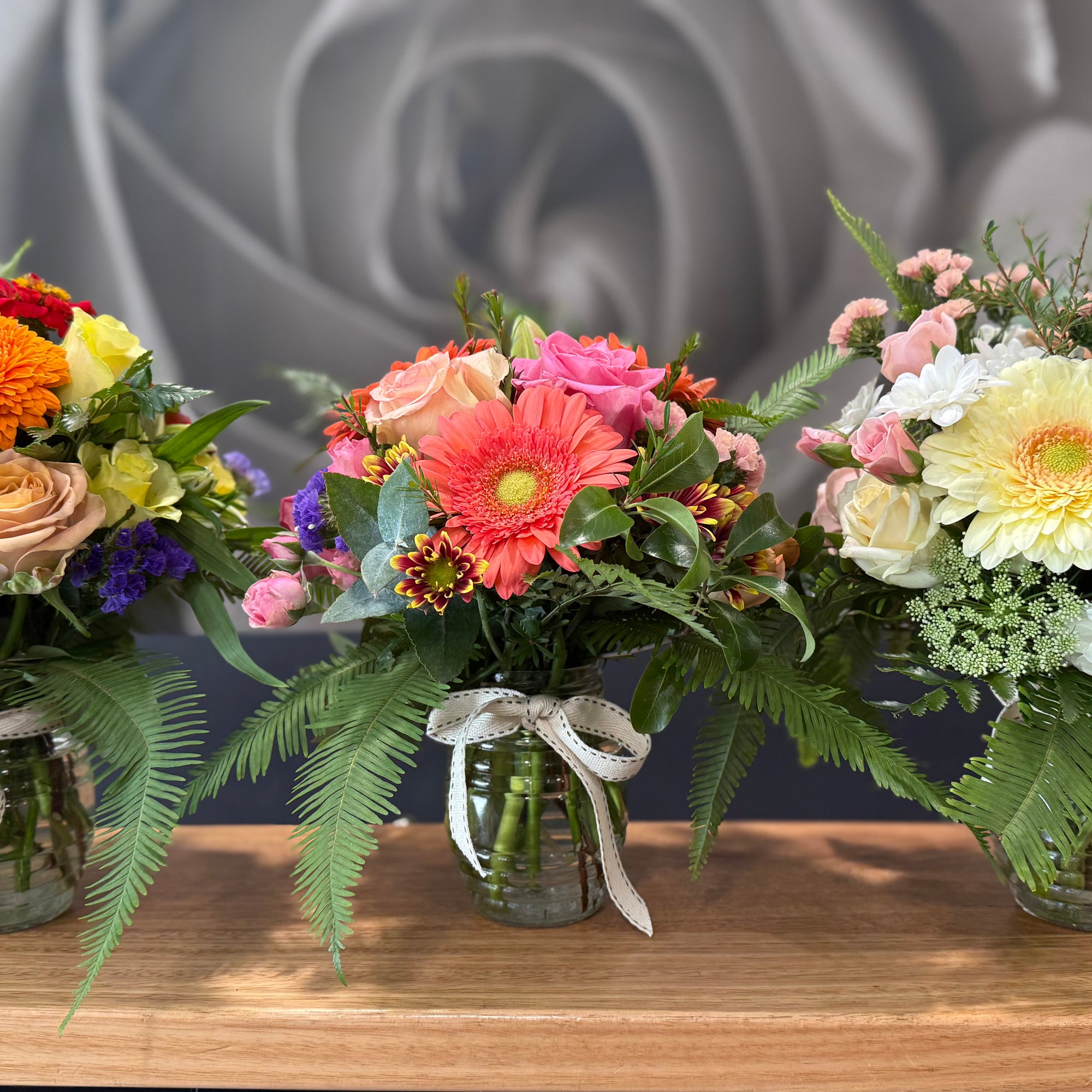 Three  small vibrant flower bouquets in clear glass vases with natural ribbon bows, displayed on a wooden table against a gray floral wall backdrop.
