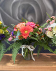 Three  small vibrant flower bouquets in clear glass vases with natural ribbon bows, displayed on a wooden table against a gray floral wall backdrop.