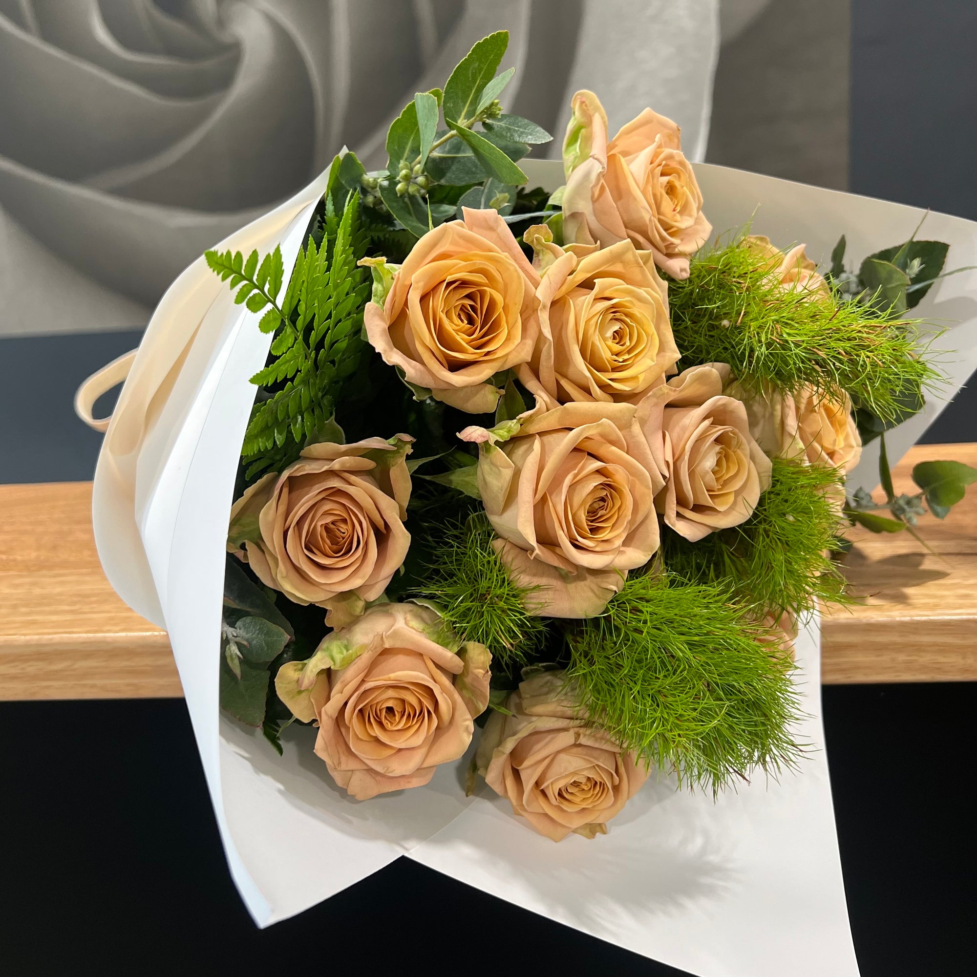 Bouquet of beige roses with greenery on a white paper wrap, placed on a wooden surface.