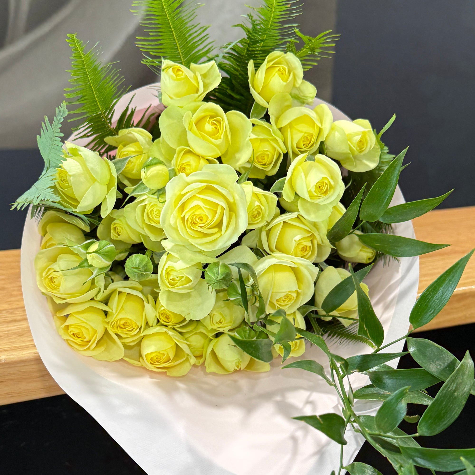 A bouquet of pale yellow spray  roses wrapped in white paper, with green foliage accents, photographed on a wooden surface against a dark background.