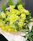 A bouquet of pale yellow spray  roses wrapped in white paper, with green foliage accents, photographed on a wooden surface against a dark background.
