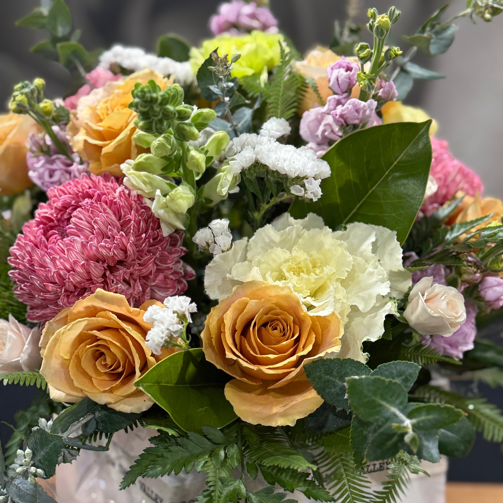 A close-up of a mixed floral box arrangement featuring yellow and peach roses, pink chrysanthemums, white carnations, lavender sprigs, and green foliage in a white vase.