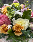 A close-up of a mixed floral box arrangement featuring yellow and peach roses, pink chrysanthemums, white carnations, lavender sprigs, and green foliage in a white vase.