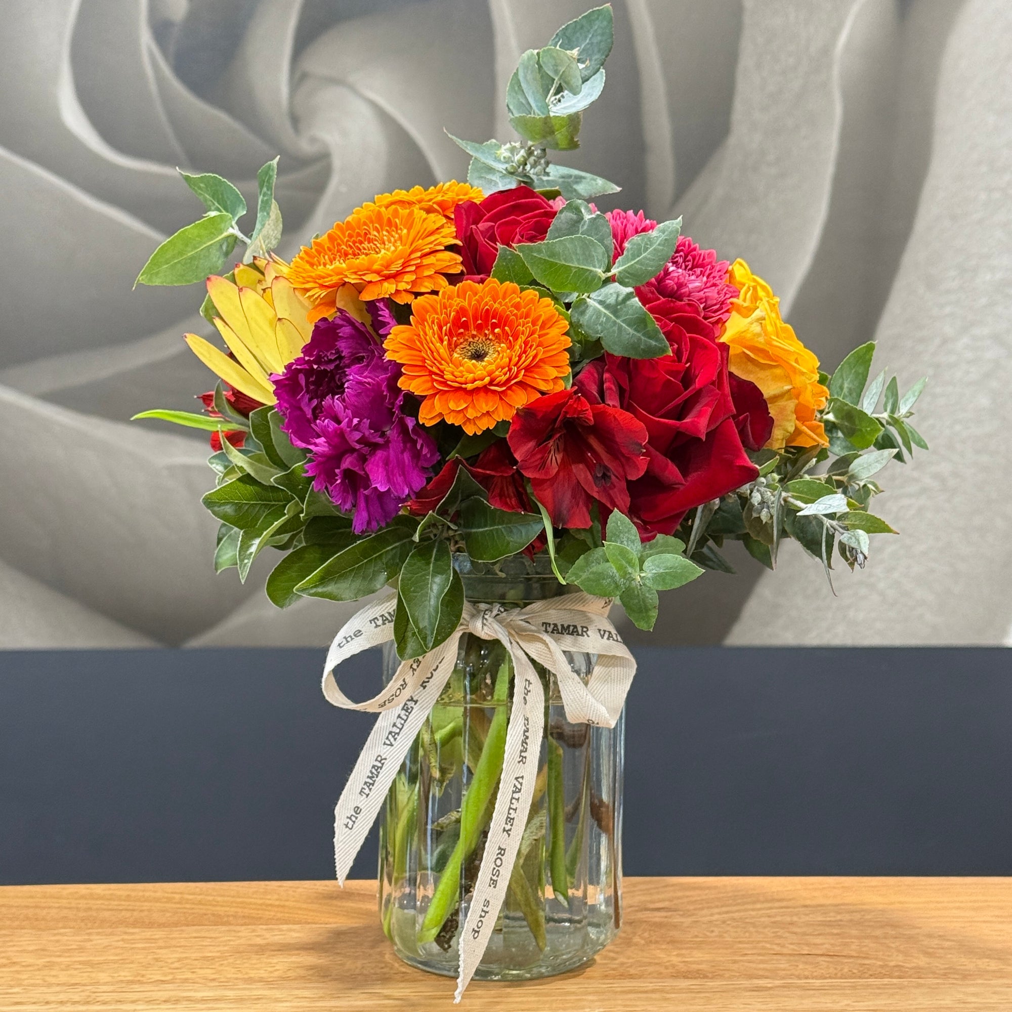 A vibrant mixed flower bouquet in a clear glass jar vase with a cream ribbon printed with 'The TAMAR VALLEY ROSE Shop', displayed on a wooden table against a gray and dark background.
