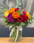 A vibrant mixed flower bouquet in a clear glass jar vase with a cream ribbon printed with 'The TAMAR VALLEY ROSE Shop', displayed on a wooden table against a gray and dark background.