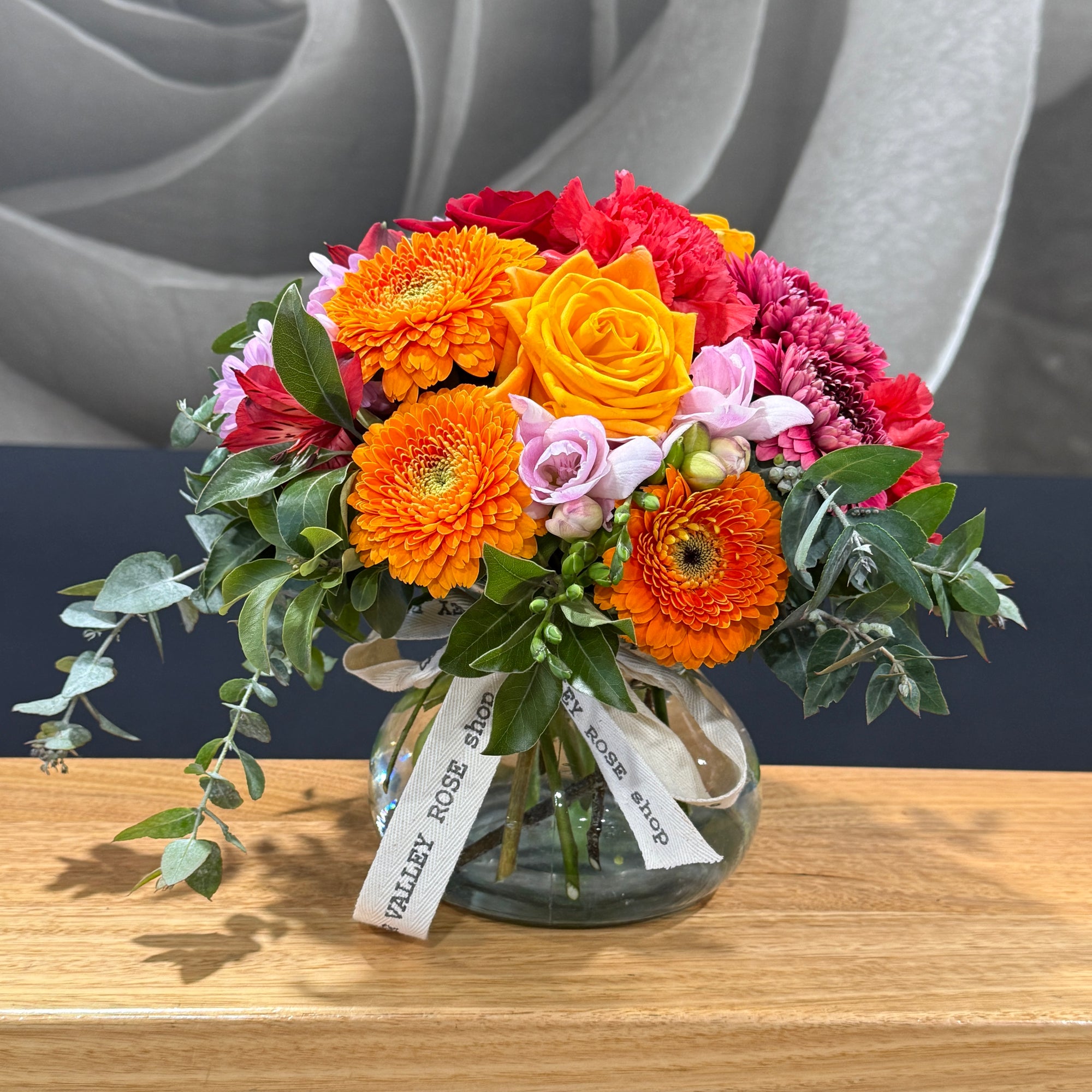 A vibrant mixed floral arrangement in a clear glass vase, featuring orange gerbera daisies, a yellow-orange rose, red and pink chrysanthemums, and green foliage, tied with a white 'Tamar Valley ROSE SHOP' ribbon, set on a wooden table.