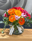 A vibrant mixed floral arrangement in a clear glass vase, featuring orange gerbera daisies, a yellow-orange rose, red and pink chrysanthemums, and green foliage, tied with a white 'Tamar Valley ROSE SHOP' ribbon, set on a wooden table.