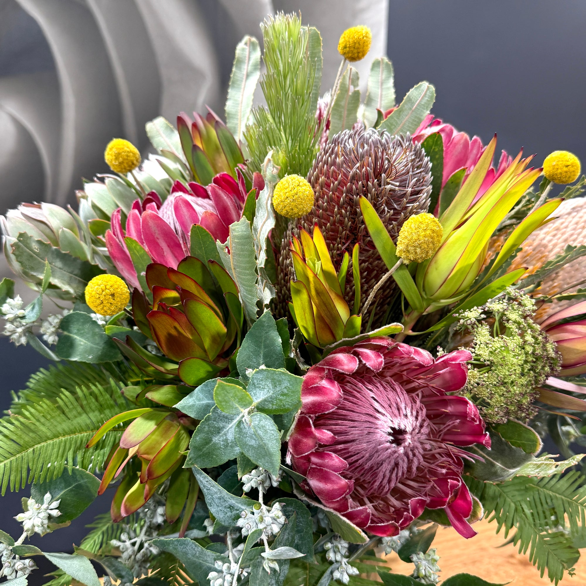A close-up of a vibrant mixed floral arrangement featuring pink protea flowers, yellow globe flowers, green and silver foliage, and textured seed pods.