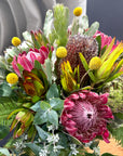 A close-up of a vibrant mixed floral arrangement featuring pink protea flowers, yellow globe flowers, green and silver foliage, and textured seed pods.