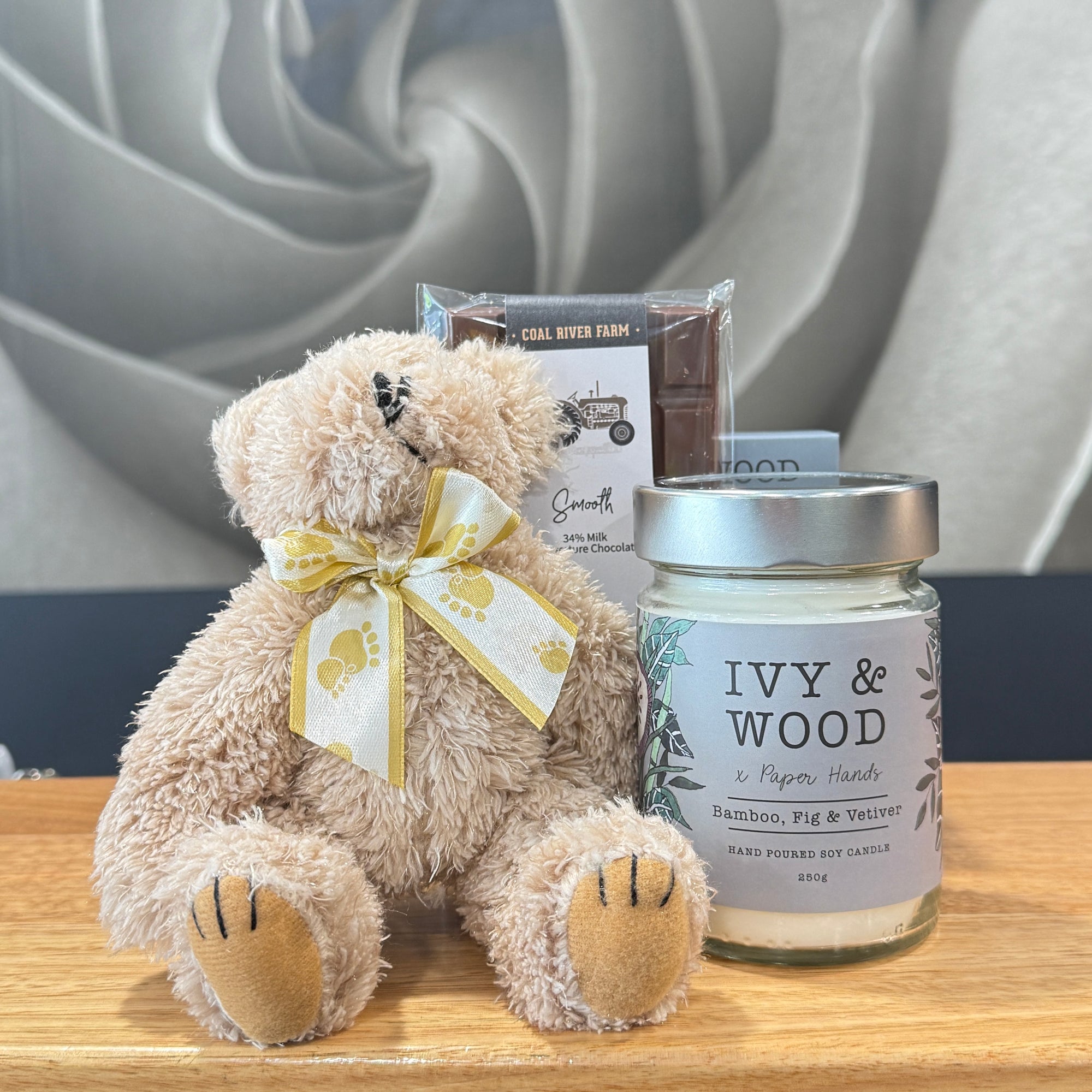 Teddy bear with a bow and a candle labeled 'Ivy & Wood'  and Chocolate Bar on a wooden surface.