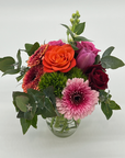 A small clear Honey Pot style jar filled with an arrangement of seasonal mixed flowers, including shades of pink, red, and green, with leaves and stems visible.