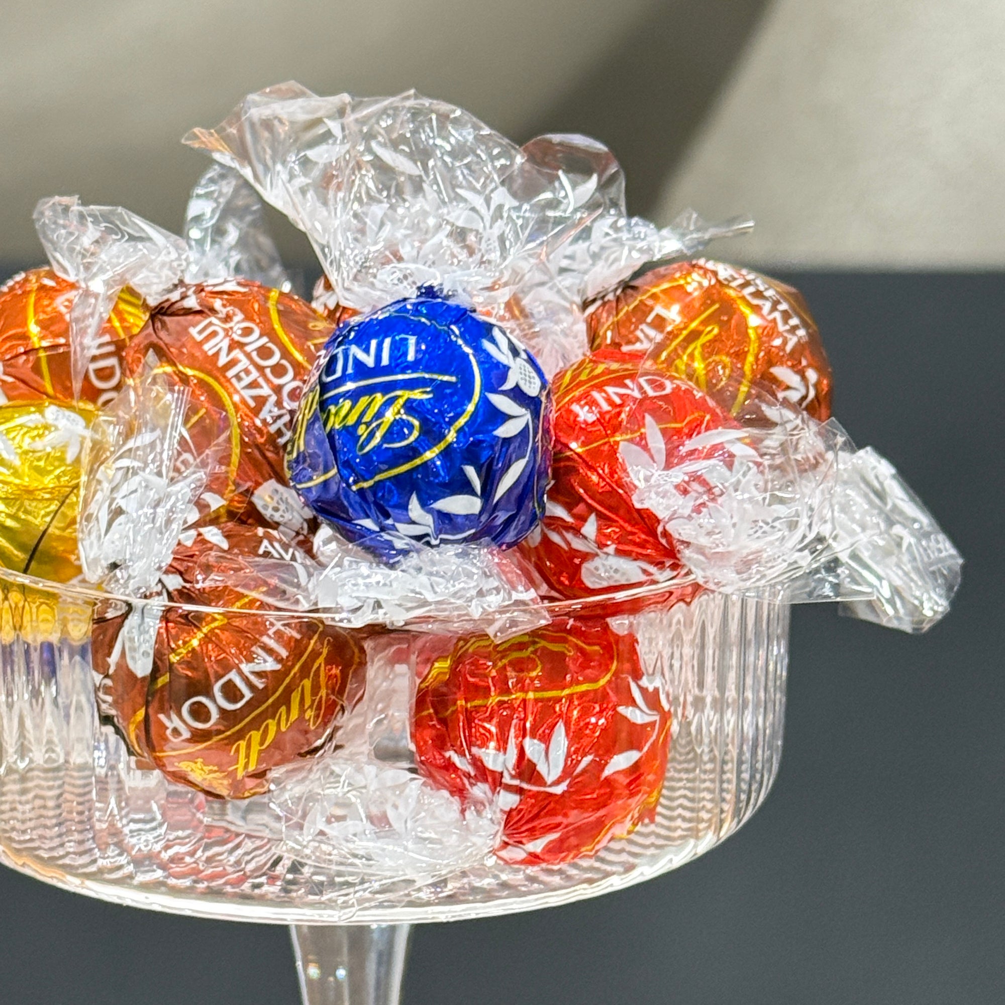 A clear glass bowl filled with assorted wrapped chocolates, including a prominent blue Lindt ball and several red, orange, and gold wrapped candies, photographed close-up against a dark gray background.