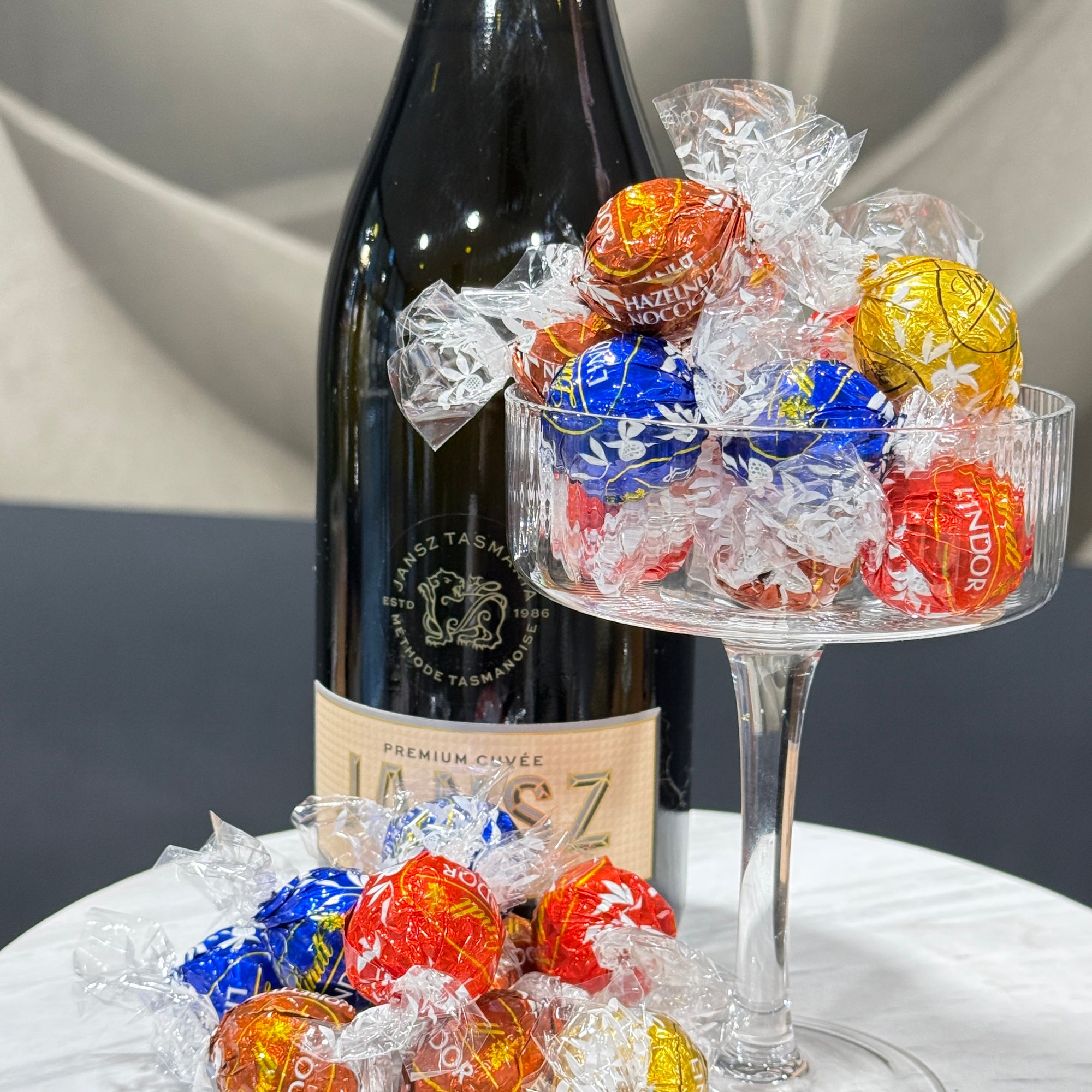 A bottle of Jansz premium cuvée sparkling wine with a clear glass pedestal bowl filled with wrapped chocolates in red, blue, yellow, and orange, Lindt Lindor with more chocolates scattered on the table in front.
