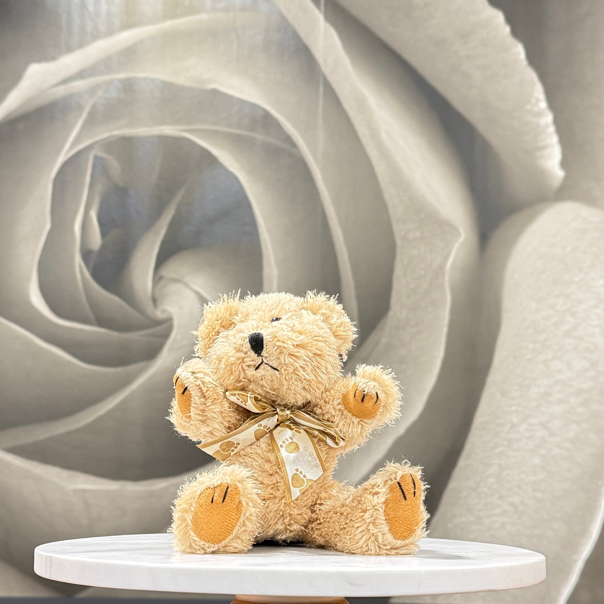 A small beige teddy bear plush toy with a gold ribbon bow sits centered on a round white pedestal against a soft gray rose-shaped backdrop.
