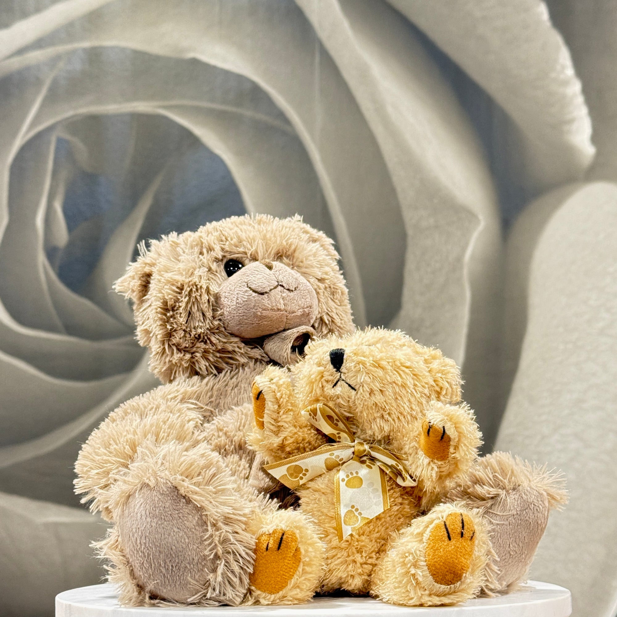 Two plush teddy bears posed together on a white pedestal. The larger bear is light tan with darker tan paws and snout, while the smaller bear is cream with yellow accents on its paws and a cream ribbon with small yellow dots.