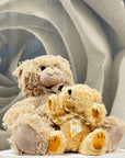Two plush teddy bears posed together on a white pedestal. The larger bear is light tan with darker tan paws and snout, while the smaller bear is cream with yellow accents on its paws and a cream ribbon with small yellow dots.