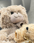 A close-up of a soft, shaggy beige teddy bear holding a smaller cream teddy bear with a ribbon bow, set against a gray upholstered background.
