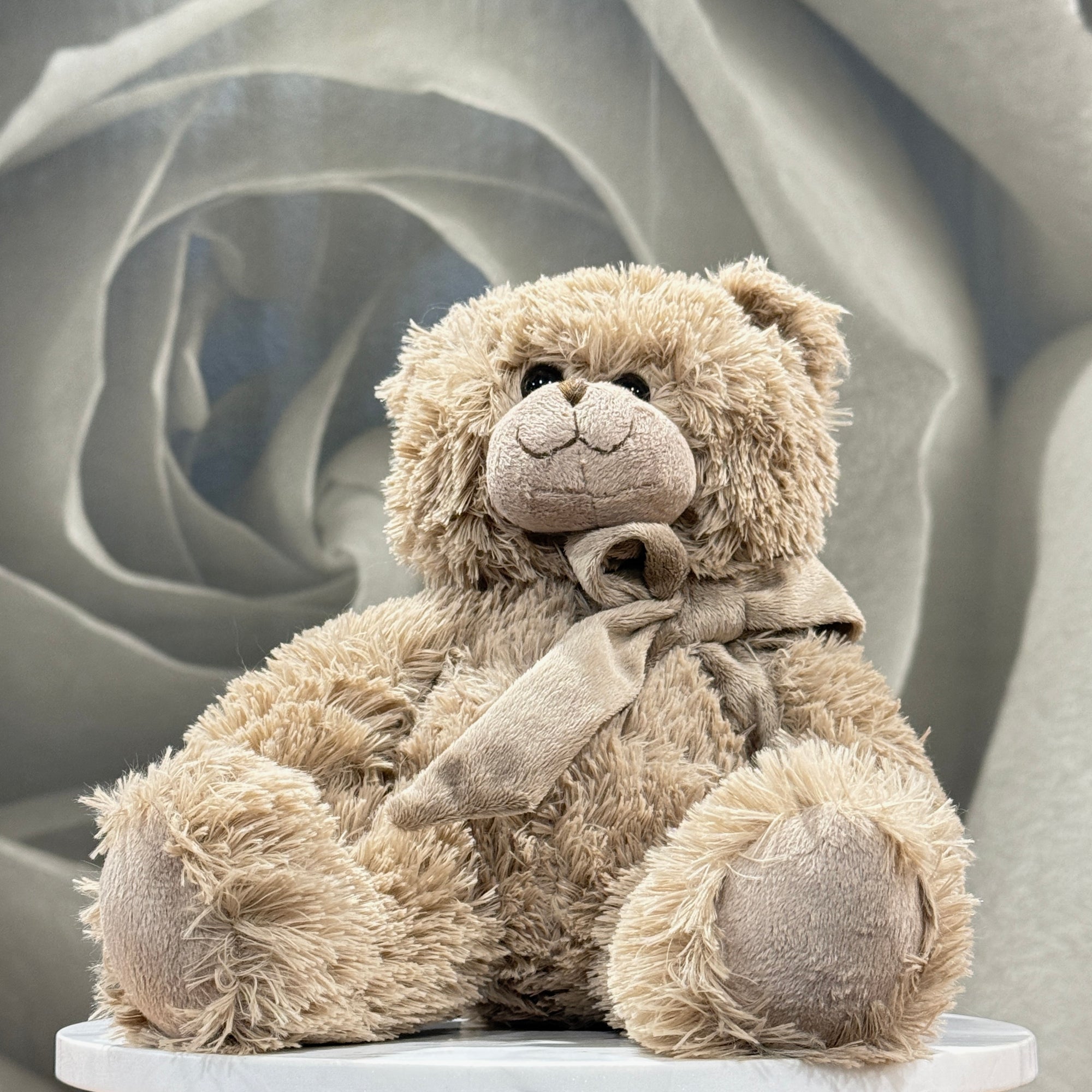A plush teddy bear in a warm beige/tan color with a matching fabric bow, sitting on a white round pedestal against a soft gray rose-shaped backdrop.
