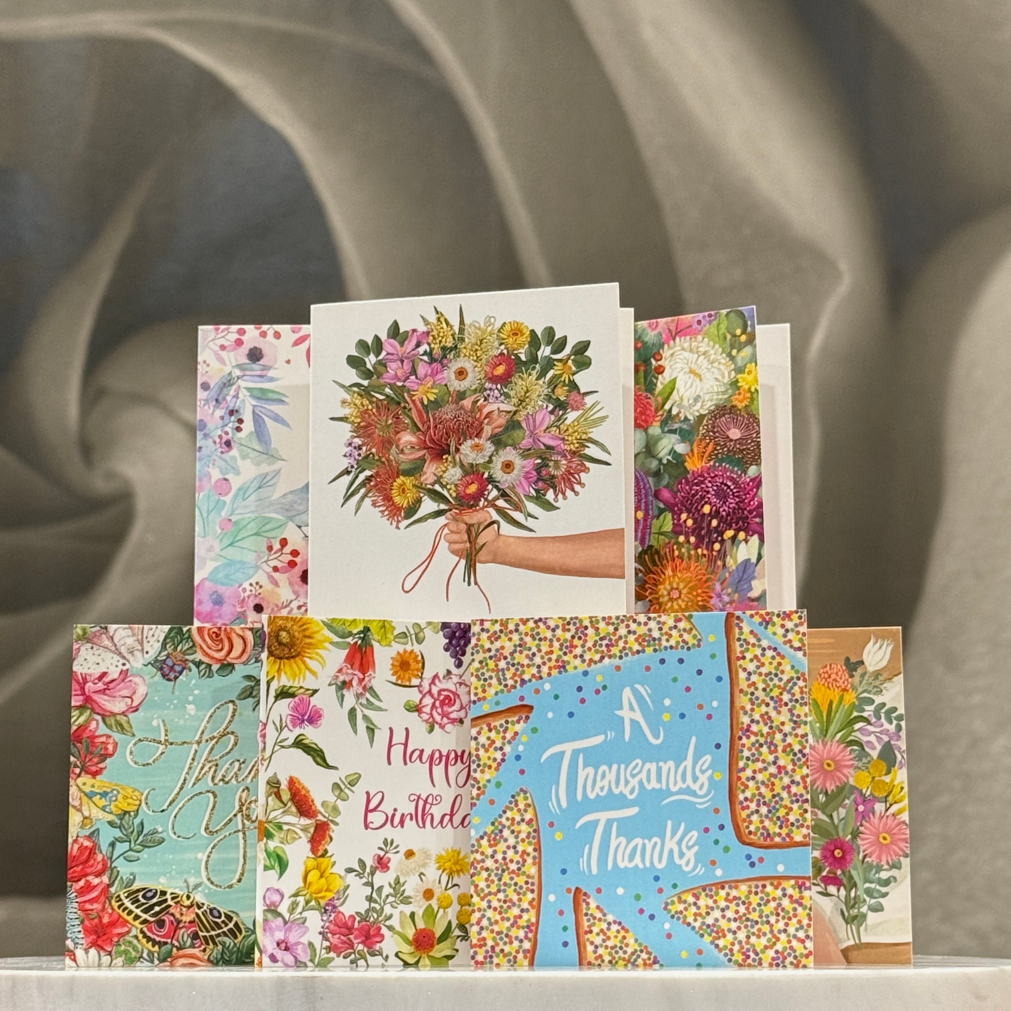 A stack of Mini La La Land floral greeting cards displayed against a draped gray fabric background. The cards feature colorful watercolor and illustration designs, including bouquets, birds, butterflies, and celebratory messages such as 'Happy Birthday' and 'A Thousand Thanks'.