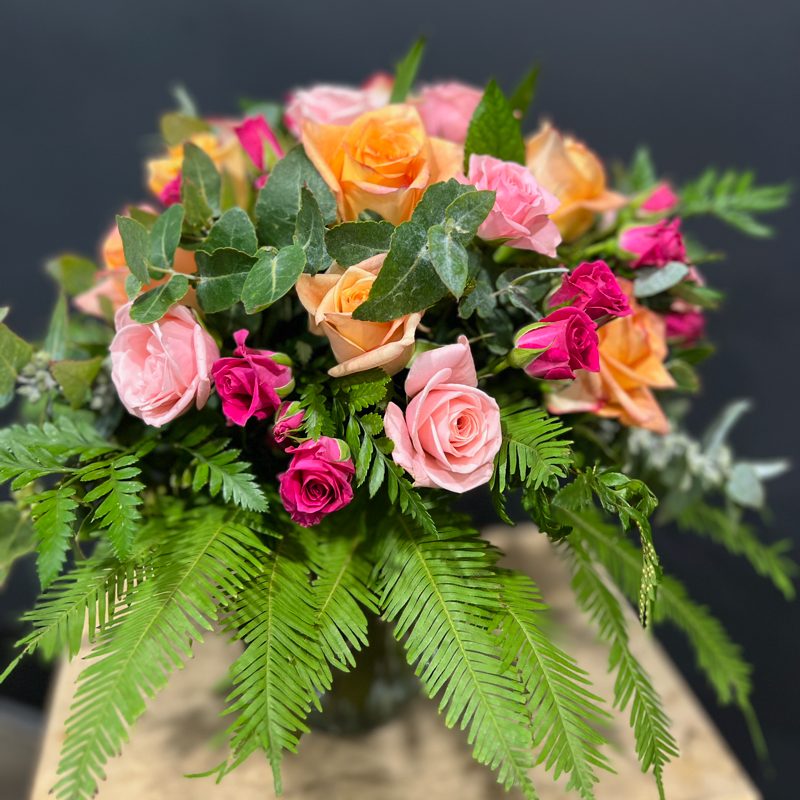 Glass Mason jar of roses, including spray roses with gum foliage and ferns on a grey background.