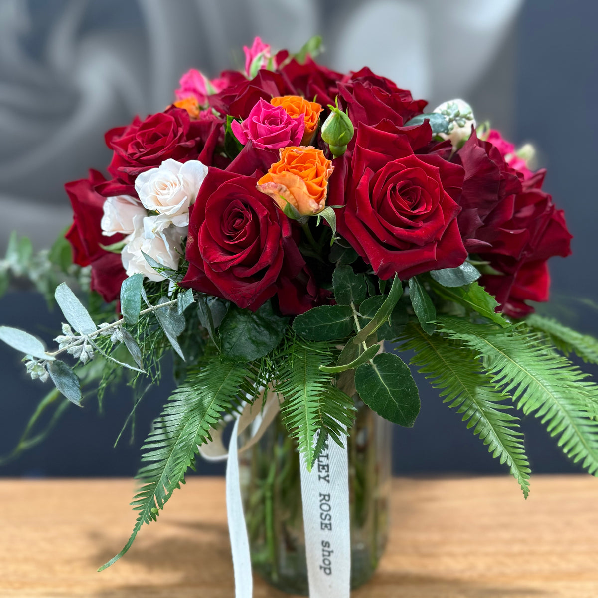 A vibrant flower arrangement featuring deep red roses, bright orange roses, white lily of the valley, and green foliage in a clear glass vase with a white ribbon tag reading 'The TAMAR VALLEY ROSE shop'.