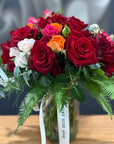 A vibrant flower arrangement featuring deep red roses, bright orange roses, white lily of the valley, and green foliage in a clear glass vase with a white ribbon tag reading 'The TAMAR VALLEY ROSE shop'.