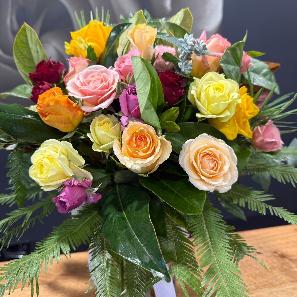 A vibrant rose bouquet featuring a mix of pastel and bright roses, yellow and orange blossoms, pink flowers, and green foliage, arranged in a mason jar container on a wooden surface.