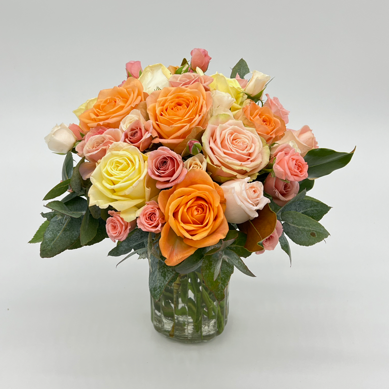 A glass Mason style jar arrangement of spray roses in shades of pink, orange, and yellow with green foliage.