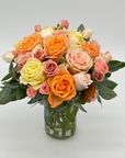 A glass Mason style jar arrangement of spray roses in shades of pink, orange, and yellow with green foliage.
