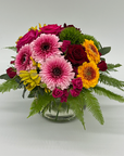A glass posy vase filled with seasonal mixed flowers including pink and yellow roses, and various foliage, placed on a neutral background.