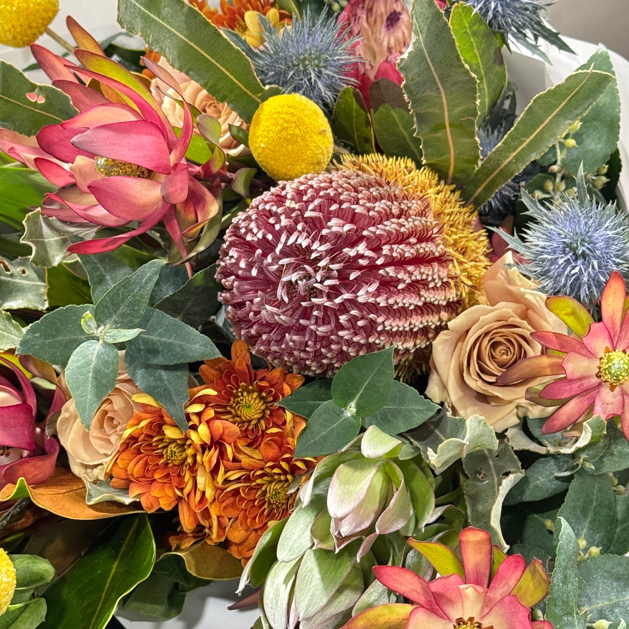A close-up of a dense floral arrangement featuring a large pink and white protea, banksia blooms, orange chrysanthemums, pale beige roses, blue sea holly, and green leaves.