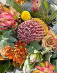 A close-up of a dense floral arrangement featuring a large pink and white protea, banksia blooms, orange chrysanthemums, pale beige roses, blue sea holly, and green leaves.