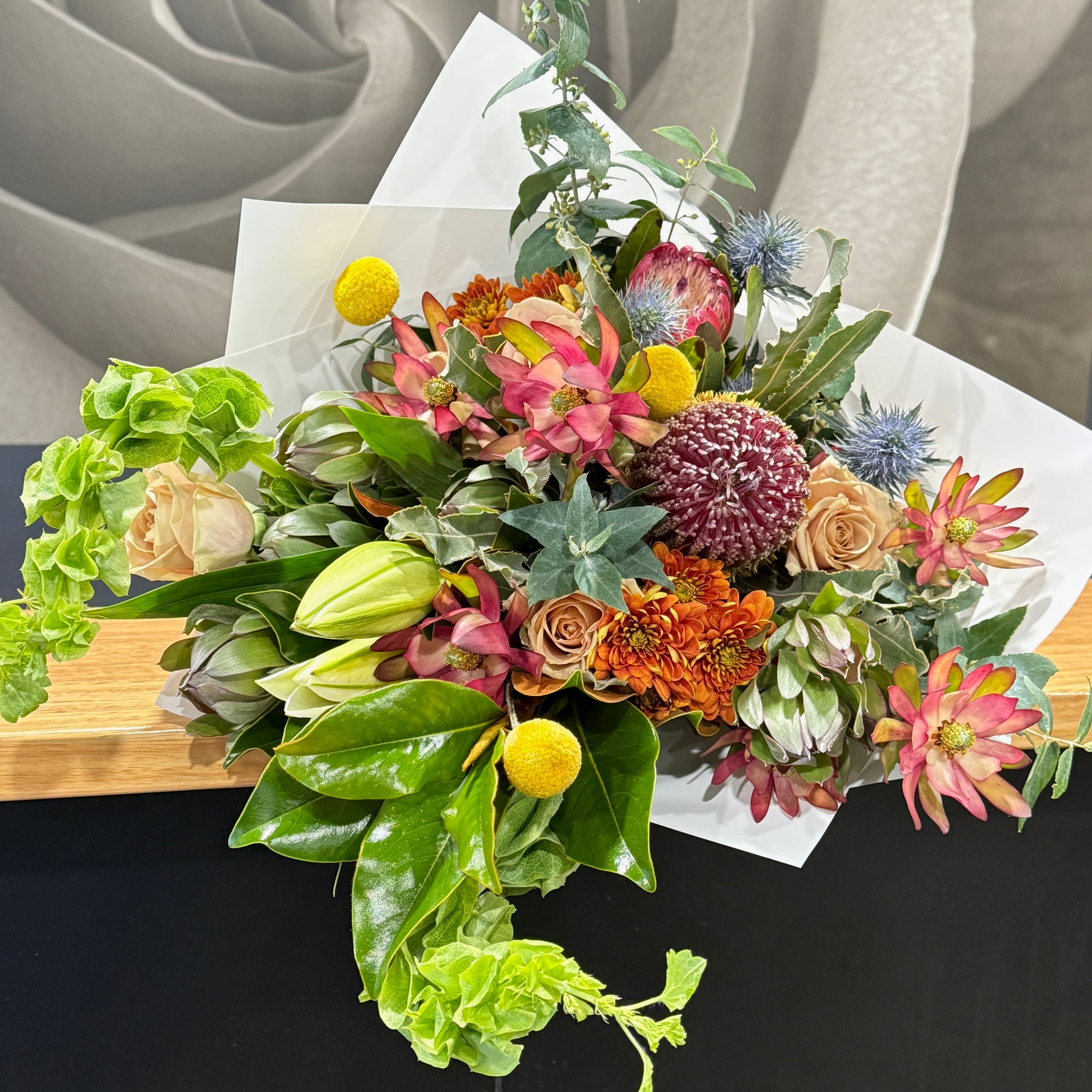A vibrant  floral arrangement featuring a mix of green leaves, gum foliage, banksia, leucodendron and orange flowers, yellow pom-pom billy buttons, and purple thistle-like blooms, styled on a wooden bench with white wrapping against a gray background.