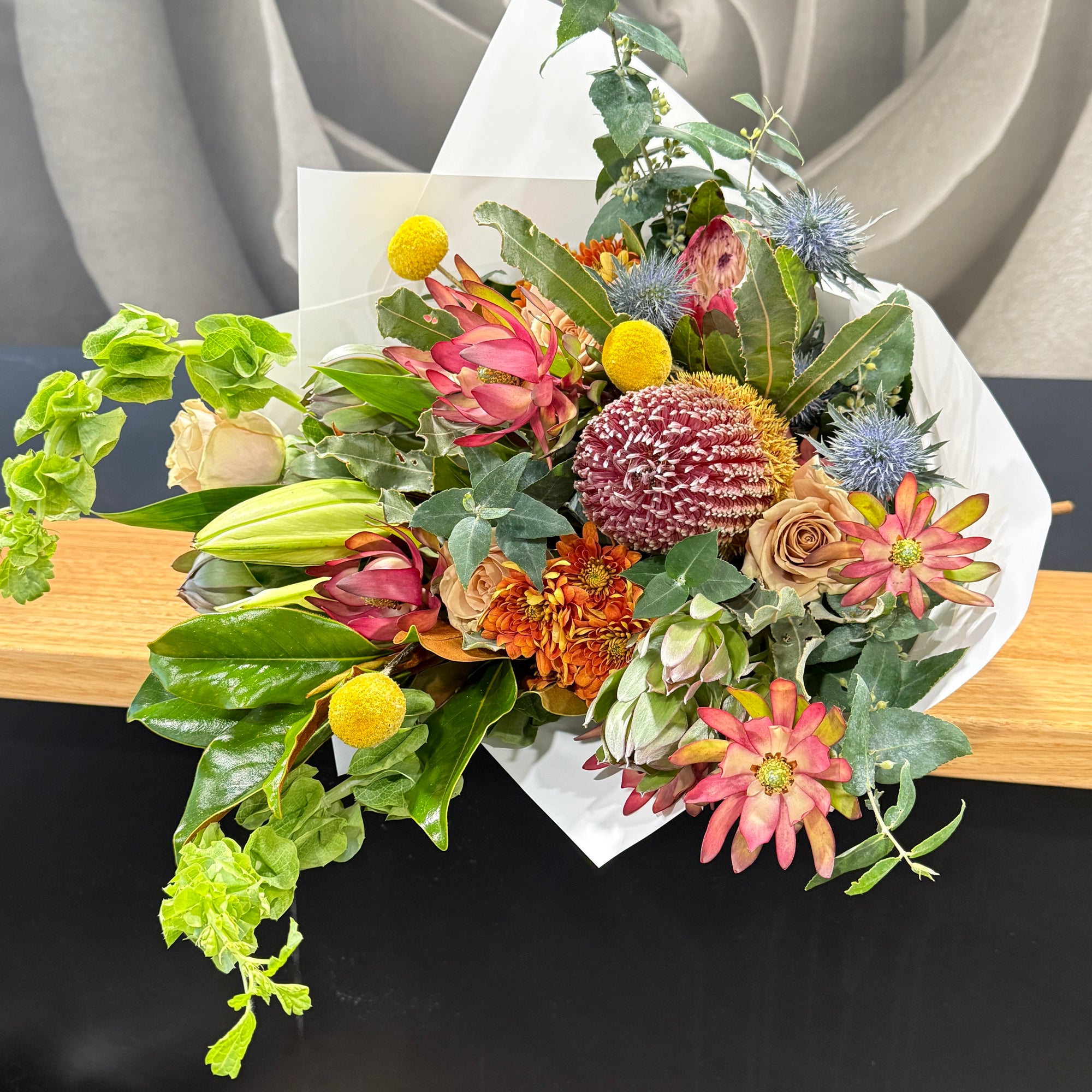 A vibrant wildflower bouquet with banksia, golden Billy Button, and green foliage, wrapped in white paper and resting on a wooden surface.
