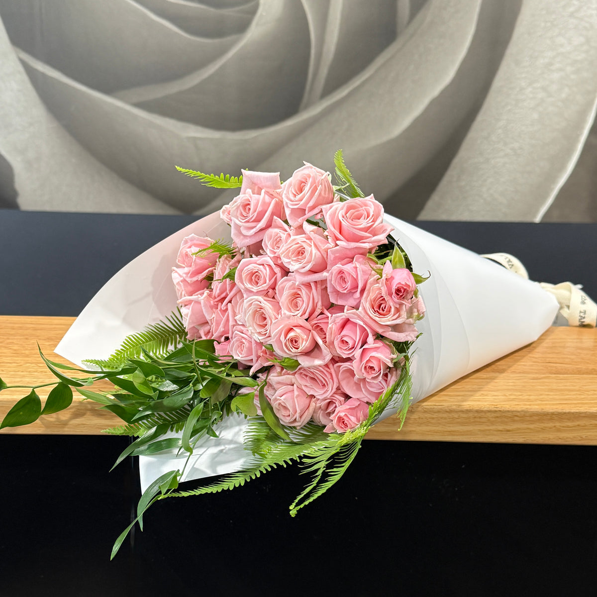 A bouquet of pink roses wrapped in white paper with green foliage, resting on a wooden surface against a dark background.
