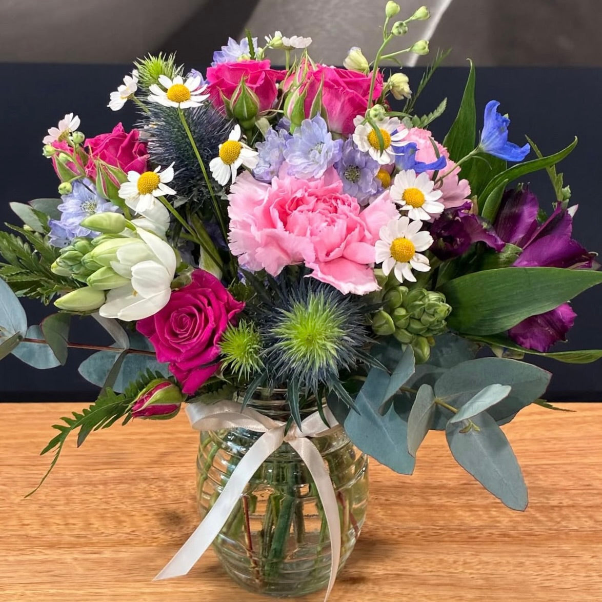 A vibrant mixed floral arrangement in a clear glass jar with a white ribbon, displayed on a wooden tabletop. The bouquet features pink carnations, pink and magenta roses, white daisies, purple flowers, blue thistles, and green foliage.
